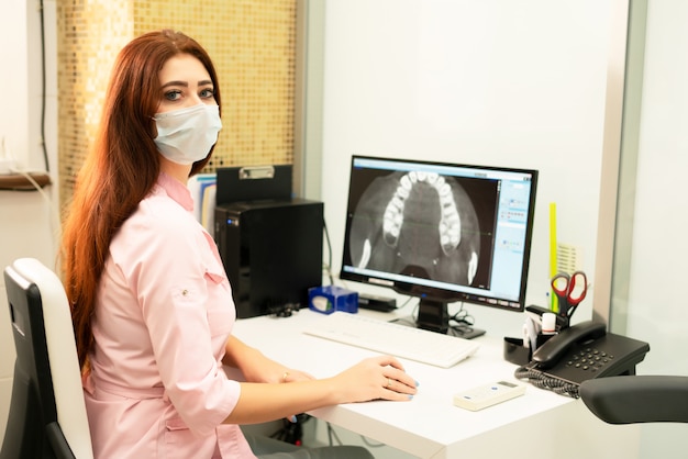 Una doctora dentista está sentada en una mesa, en una computadora, una tomografía computarizada de la mandíbula. El doctor está vestido con ropa profesional.