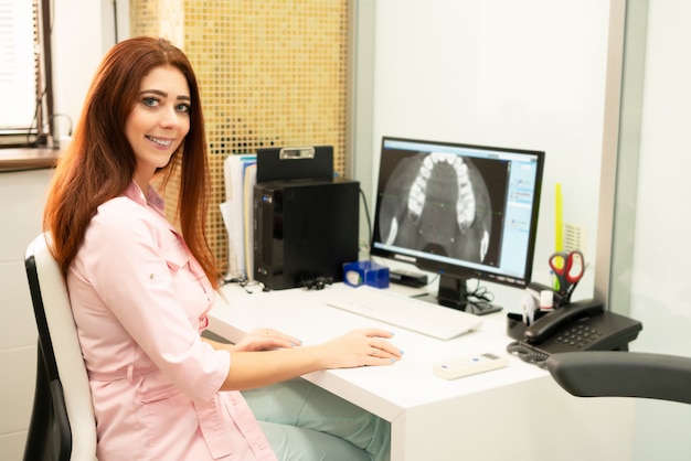 Una doctora dentista está sentada en una mesa, en una computadora, una tomografía computarizada de la mandíbula. El doctor está vestido con ropa profesional.
