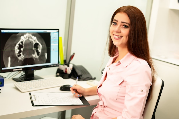 Una doctora dentista está sentada en una mesa, en una computadora, una tomografía computarizada de la mandíbula. El doctor está vestido con ropa profesional.