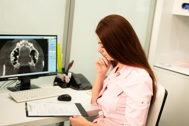 Doctora dentista está sentado a la mesa en la computadora una tomografía computarizada de la mandíbula. El doctor está vestido con ropa profesional.