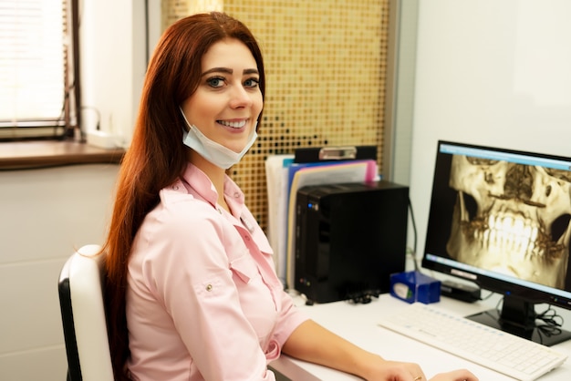 Foto una doctora dentista está sentada en una mesa, en una computadora, una tomografía computarizada de la mandíbula. el doctor está vestido con ropa profesional.