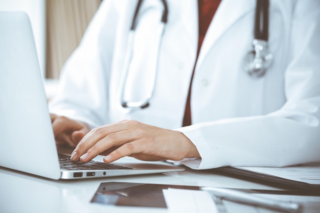 Una doctora desconocida escribiendo en una computadora portátil mientras está sentada en la mesa. Concepto de medicina.