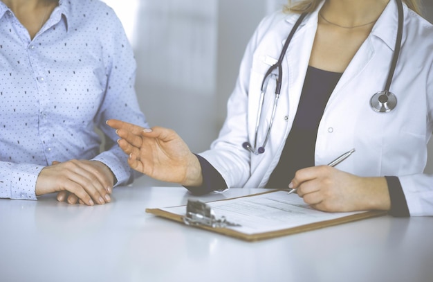 Una doctora desconocida está anotando los síntomas de un resfriado del paciente, mientras se sientan juntos en el escritorio del gabinete de una clínica. El médico está usando un portapapeles y un estetoscopio, primer plano. perfecto yo