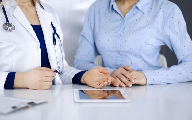 Foto una doctora desconocida le muestra a su paciente una descripción de la medicación, mientras se sientan juntas en el escritorio del gabinete de una clínica. la doctora está usando una tableta de computadora y un estetoscopio