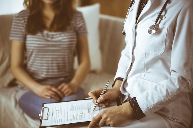 Una doctora desconocida y una paciente discutiendo el examen de salud actual mientras están sentadas en una acogedora sala de estar. Concepto de estancia en casa durante la pandemia de Coronavirus y la cuarentena de autoaislamiento