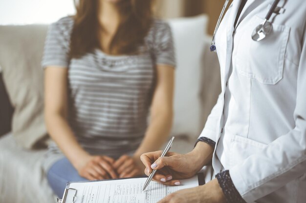 Una doctora desconocida y una paciente discutiendo el examen de salud actual mientras están sentadas en una acogedora sala de estar. Quédese en el concepto de casa durante la pandemia de Coronavirus y la cuarentena de autoaislamiento.