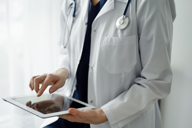 Una doctora desconocida que usa una tableta o un panel táctil mientras está de pie cerca de la ventana en la clínica. Ropa de trabajo azul oscuro de médico. concepto de medicina