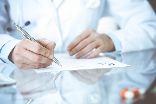 Doctora desconocida en el trabajo en el hospital. Píldoras en el portapapeles médico. Una joven médica escribe una receta o llena un formulario médico mientras está sentada en la oficina del hospital, en primer plano.