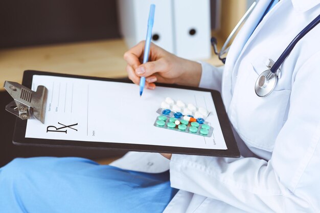 Foto doctora desconocida en el trabajo en el hospital. píldoras en el portapapeles médico. una joven médica escribe una receta o llena un formulario médico mientras está sentada en la oficina del hospital, en primer plano.