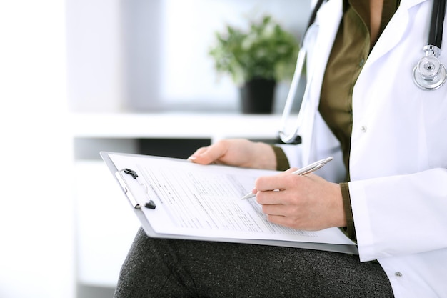 Doctora escribiendo algo en el portapapeles mientras está sentada en la silla de primer plano Terapeuta en el trabajo llenando registros de historial de medicamentos Concepto de medicina y atención médica