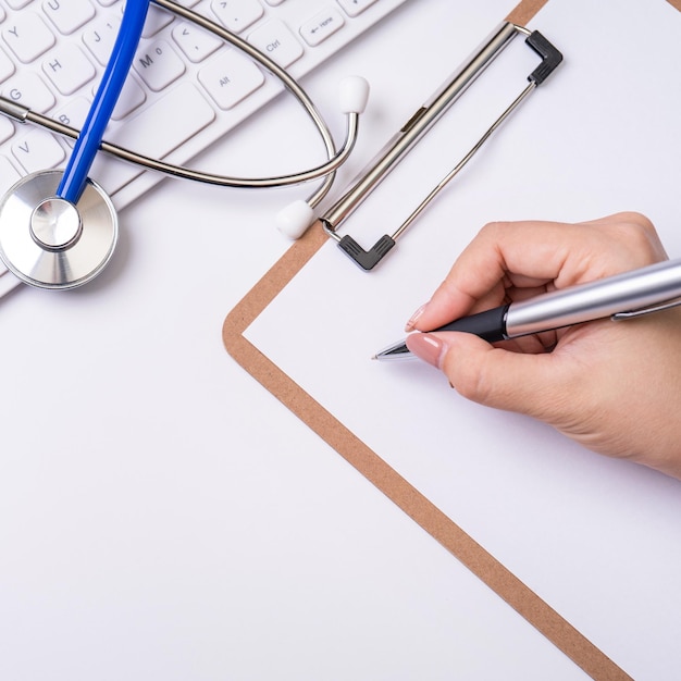 Doctora escribiendo un caso de registro médico sobre el portapapeles en la mesa de trabajo blanca con teclado de computadora estetoscopio Vista superior espacio de copia plana