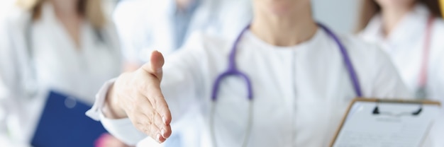 Doctora extiende su mano para saludar con el telón de fondo de colegas médicos