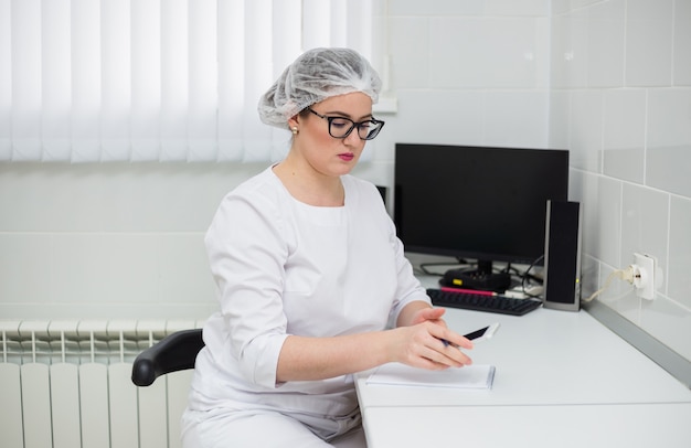 Doctora con gafas y un traje médico blanco se sienta en un escritorio con un teléfono y un cuaderno en la oficina de la clínica