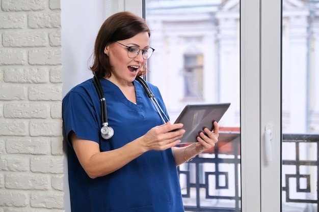 Doctora hablando en línea con el paciente mediante tableta digital, videollamada, tecnología, médico junto a la ventana en la oficina de la clínica