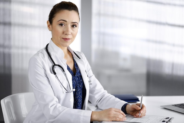 Una doctora inteligente y alegre está usando un portapapeles y escribiendo algunos nombres de medicamentos, mientras está sentada en el escritorio de su gabinete en una clínica. retrato, de, profesional, hembra, médico, con, un, stethos