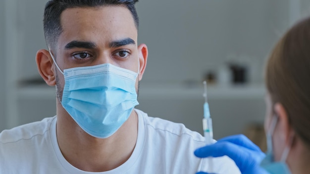 Una doctora irreconocible con guantes sostiene una jeringa con el medicamento de la vacuna preparándose para hacer una inyección de vacunación del coronavirus covid19 a un paciente árabe hispano enfermo con una máscara médica de cerca