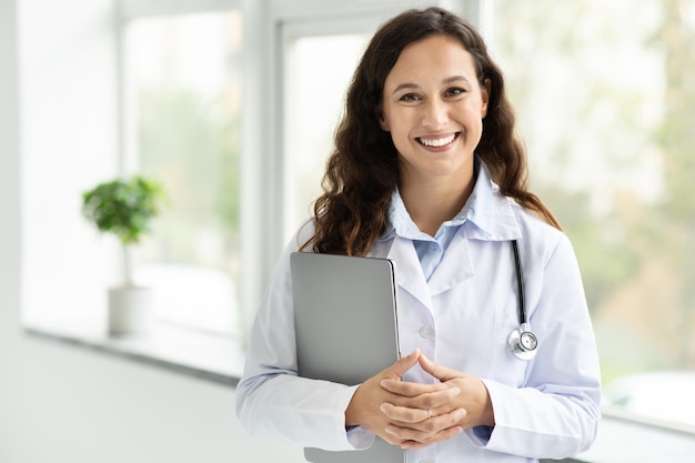 Doctora joven sonriente con computadora portátil posando en la oficina