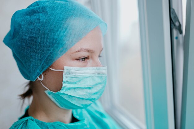 Doctora joven en uniforme y máscara mira por la depresión de la ventana