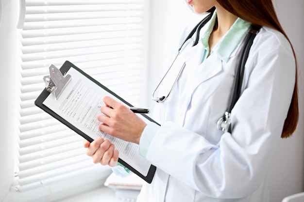 Doctora llenando el historial médico de pie cerca de la ventana en el hospital.