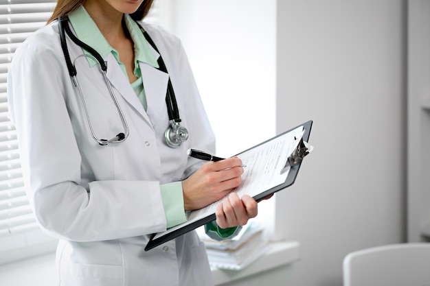 Doctora llenando el historial médico de pie cerca de la ventana en el hospital.