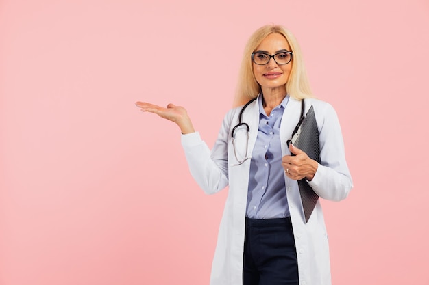 Doctora madura con gafas con estetoscopio sosteniendo una carpeta y mostrando algo en el espacio de la copia el fondo rosa