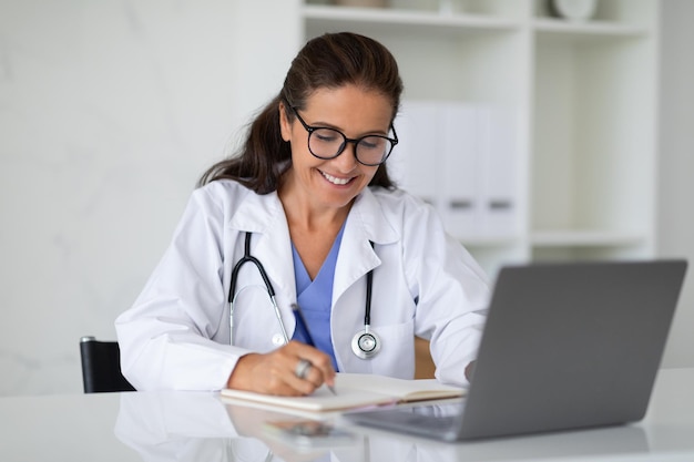 Foto doctora madura sentada en un escritorio con una computadora portátil tomando notas