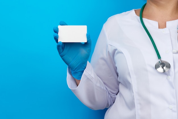 Foto doctora mano en guantes médicos de látex azul tiene una tarjeta de papel vacía sobre un fondo azul.