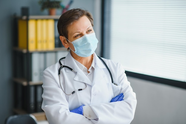 Doctora mayor con mascarilla y abrigo médico blanco de pie en el retrato del hospital