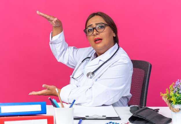 Doctora de mediana edad en bata blanca con estetoscopio mirando mostrando gesto de tamaño con las manos sentado en la mesa con carpetas de oficina sobre fondo rosa