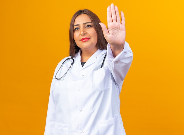 Doctora de mediana edad en bata blanca con estetoscopio con rostro serio haciendo gesto de parada con la mano de pie sobre la pared naranja