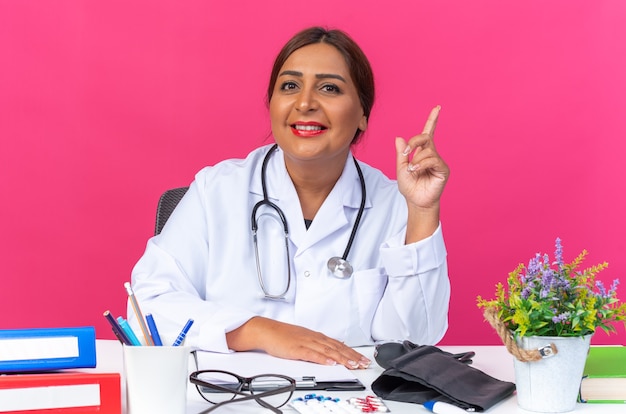 Doctora de mediana edad en bata blanca con estetoscopio sonriendo confiado mostrando el dedo índice con nueva idea sentado en la mesa con carpetas de oficina en rosa