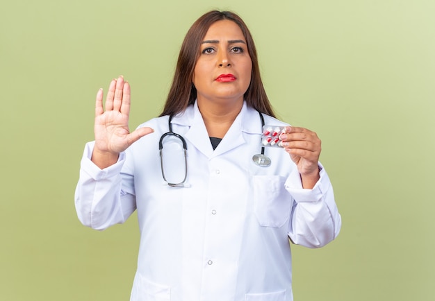 Doctora de mediana edad en bata blanca con estetoscopio sosteniendo blister con pastillas mirando al frente con cara seria mostrando la mano abierta de pie sobre la pared verde