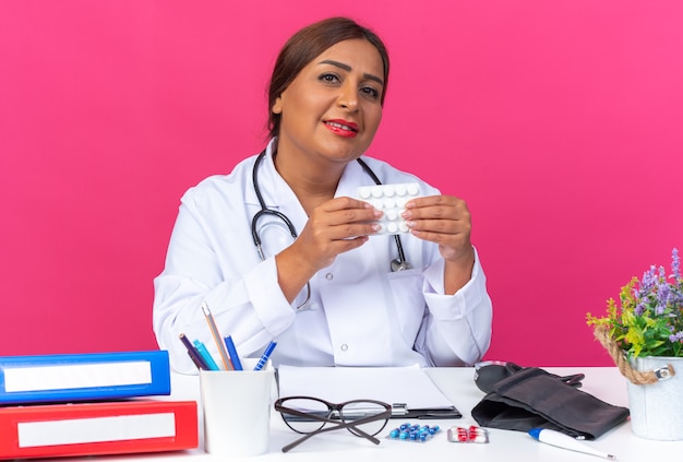 Doctora de mediana edad en bata blanca con estetoscopio sosteniendo blister con pastillas mirando con sonrisa en la cara sentado en la mesa con carpetas de oficina sobre fondo rosa