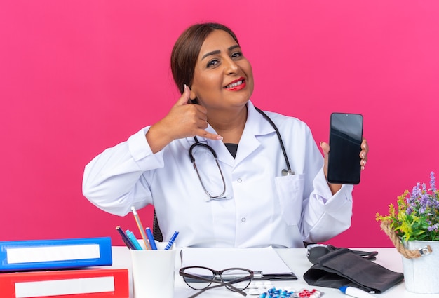 Foto doctora de mediana edad en bata blanca con estetoscopio sosteniendo teléfono inteligente haciendo gesto de llamarme sonriendo alegremente sentado en la mesa con carpetas de oficina en rosa