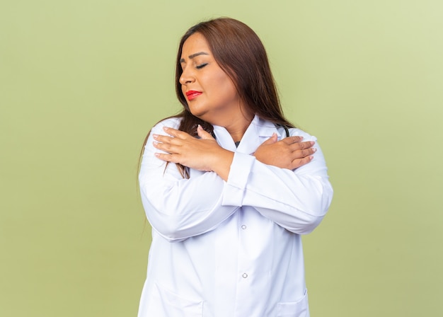 Doctora de mediana edad en bata blanca con estetoscopio tomados de la mano en el pecho con los ojos cerrados sintiendo emociones positivas de pie sobre la pared verde