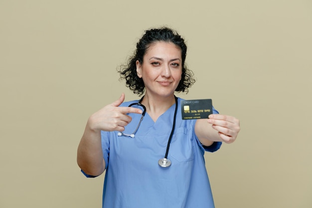 Doctora de mediana edad confiada y complacida con uniforme y estetoscopio alrededor del cuello estirando la tarjeta de crédito hacia la cámara apuntándola mirando la cámara aislada en el fondo verde oliva