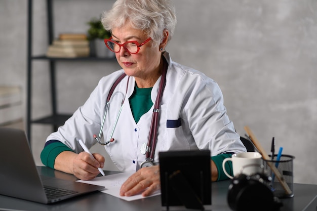 Doctora de mediana edad usando una computadora portátil escribiendo notas con un estetoscopio en la mesa concepto de atención médica...