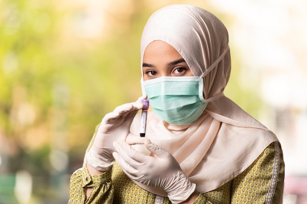 Doctora musulmana con máscara quirúrgica y guantes sosteniendo un tubo de análisis de sangre que está firmado como Covid-19