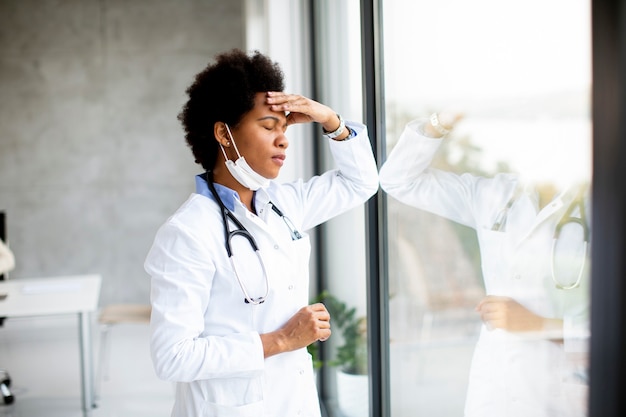 Doctora negra de pie junto a la ventana del consultorio médico con expresión negativa