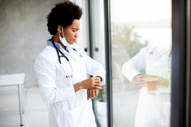 Doctora negra de pie junto a la ventana de la oficina médica y comprobando el tiempo de servicio