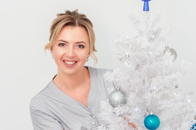 Doctora o cosmetóloga de pie y sonriendo cerca del árbol de Navidad en la pared blanca
