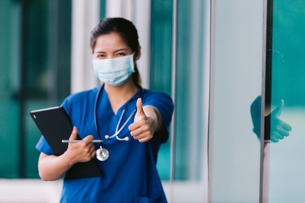 Doctora o enfermera asiática con mascarilla sosteniendo portapapeles y mostrando los pulgares hacia arriba en el hospital