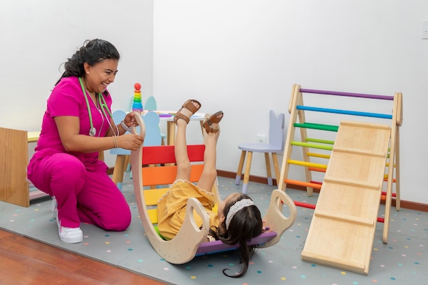 Doctora pediátrica latina riendo mientras juega con una niña en el área de juegos de su oficina