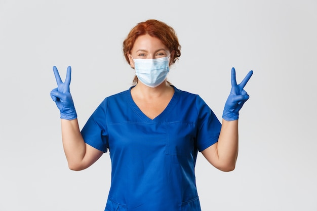 Doctora pelirroja sonriente optimista, médico con máscara médica y guantes mostrando gesto de paz, manteniéndose positivo durante la cuarentena.