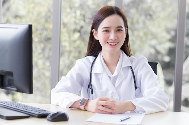 Una doctora profesional asiática sentada sonríe con una túnica blanca y un estetoscopio mientras espera