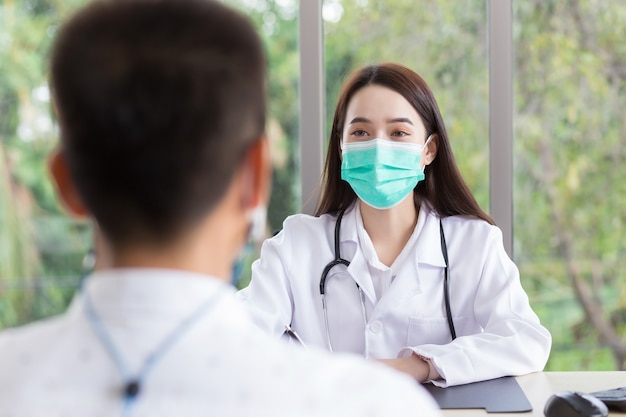 La doctora profesional asiática usa bata médica y mascarilla mientras examina y habla con un paciente en la sala de examen en el hospital.