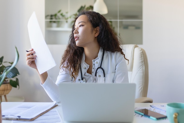 Doctora profesional médica asiática vistiendo uniforme tomando notas en un diario médico llenando documentos historial de enfermedades del paciente mirando la pantalla de una computadora portátil estudiante viendo un seminario web