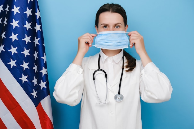 Doctora profesional poniéndose mascarilla médica protectora, viste uniforme y estetoscopio, posando sobre fondo azul con la bandera de Estados Unidos. Covid 19, trabajadores de la salud y concepto de prevención de virus