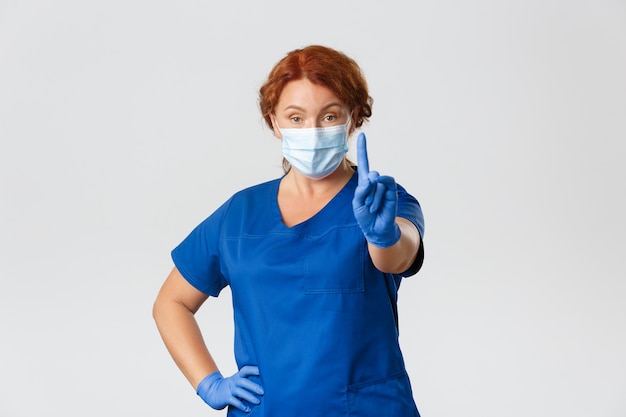 Doctora profesional seria, enfermera con mascarilla y guantes que advierten a las personas, agitando el dedo en la prohibición de restricción.