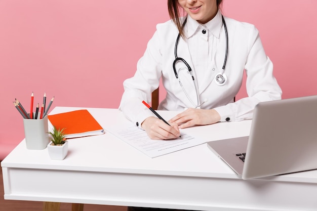 Doctora recortada emplazamiento en el escritorio trabajando llenando documentos médicos en el hospital aislado sobre fondo de pared rosa pastel en el interior. Mujer en estetoscopio de bata médica. Concepto de medicina sanitaria.